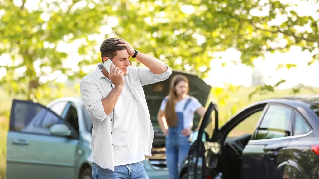 A worried man on the phone after a car accident, with a woman standing near a damaged vehicle.